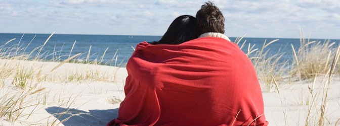 Paar kuschelt sich am Strand in Decke und blickt zum Horizont
